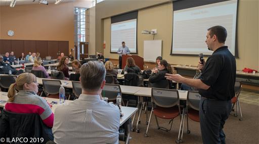 Man holding microphone stands speaking to the right of the seated attendees.